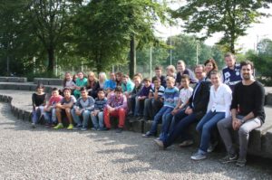 Eine Gruppe von Schülerinnen und Schülern sitzt und steht auf einer Stufe vor einer Mauer. Im Hintergrund sind Bäume und ein Zaun zu sehen.