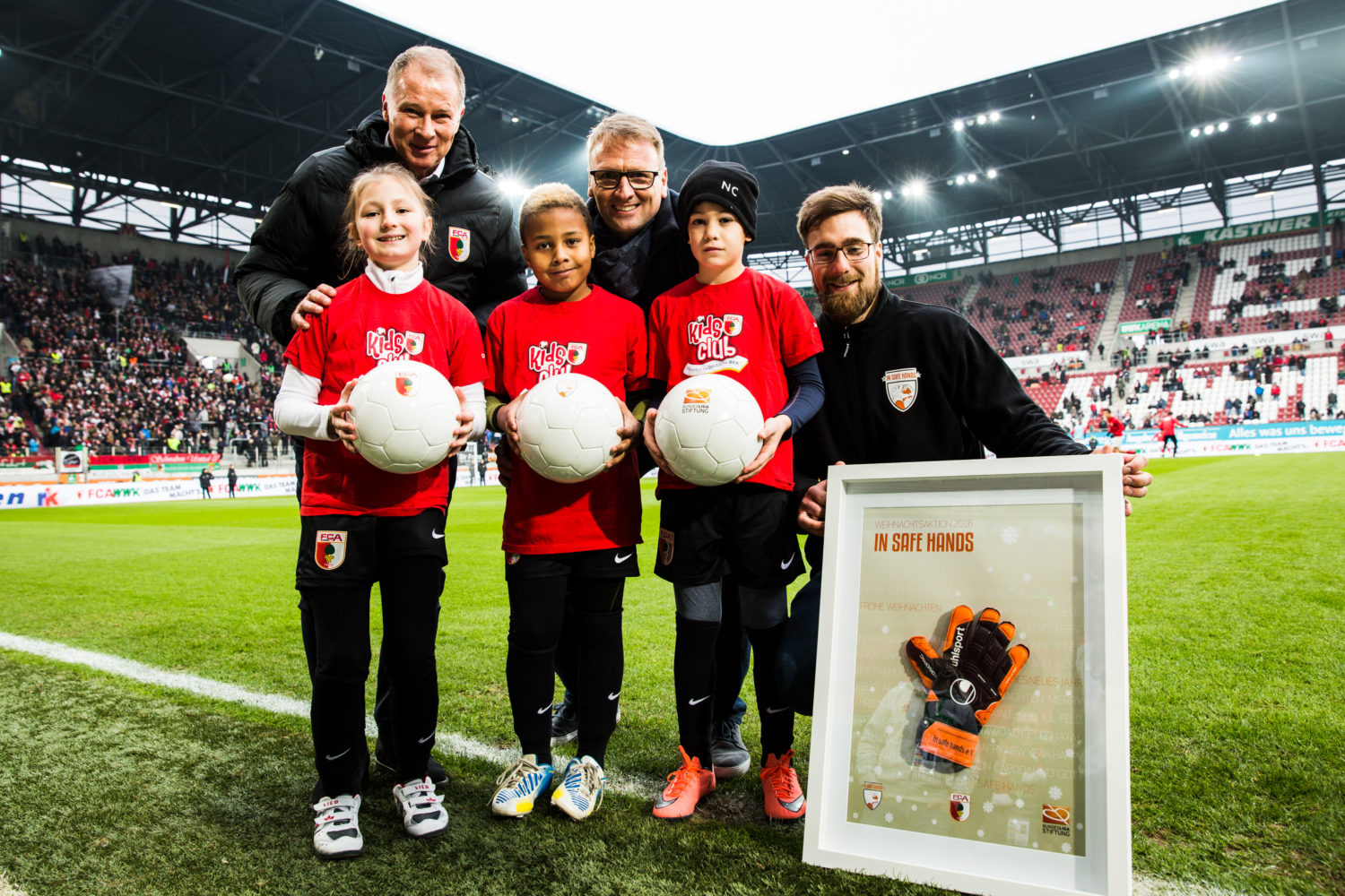 Stefan Reuter (Geschäftsführer Sport des FC Augsburg), Stefan Kiefer (Vorstandsvorsitzender der DFL Stiftung) und Andreas Luthe (Torwart des FC Augsburg) stehen mit 3 Kindern, die jeweils einen Ball in der Hand halten, im Augsburger Stadion. Andreas Luthe hält dabei einen Bilderrahmen in dem Torwarthandschuhe und die Aufschrift "in safe hands" zu sehen ist.