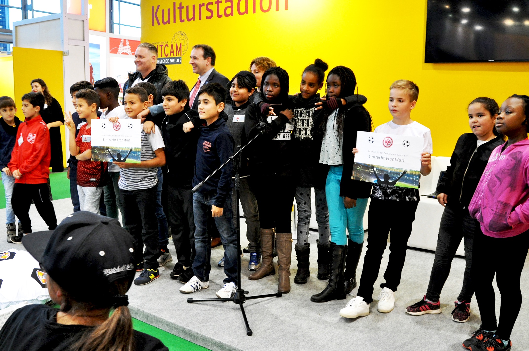 Eine Gruppe von Kindern und Jugendlichen steht im Kulturstadion und hält Bücher mit dem Covermotiv "Frankfurt Graffiti" in den Händen. Einige der Kinder schauen in die Kamera, andere blicken auf die Bücher. Im Hintergrund sind weitere Personen und ein Banner mit dem Schriftzug "Kulturstadion" zu sehen.