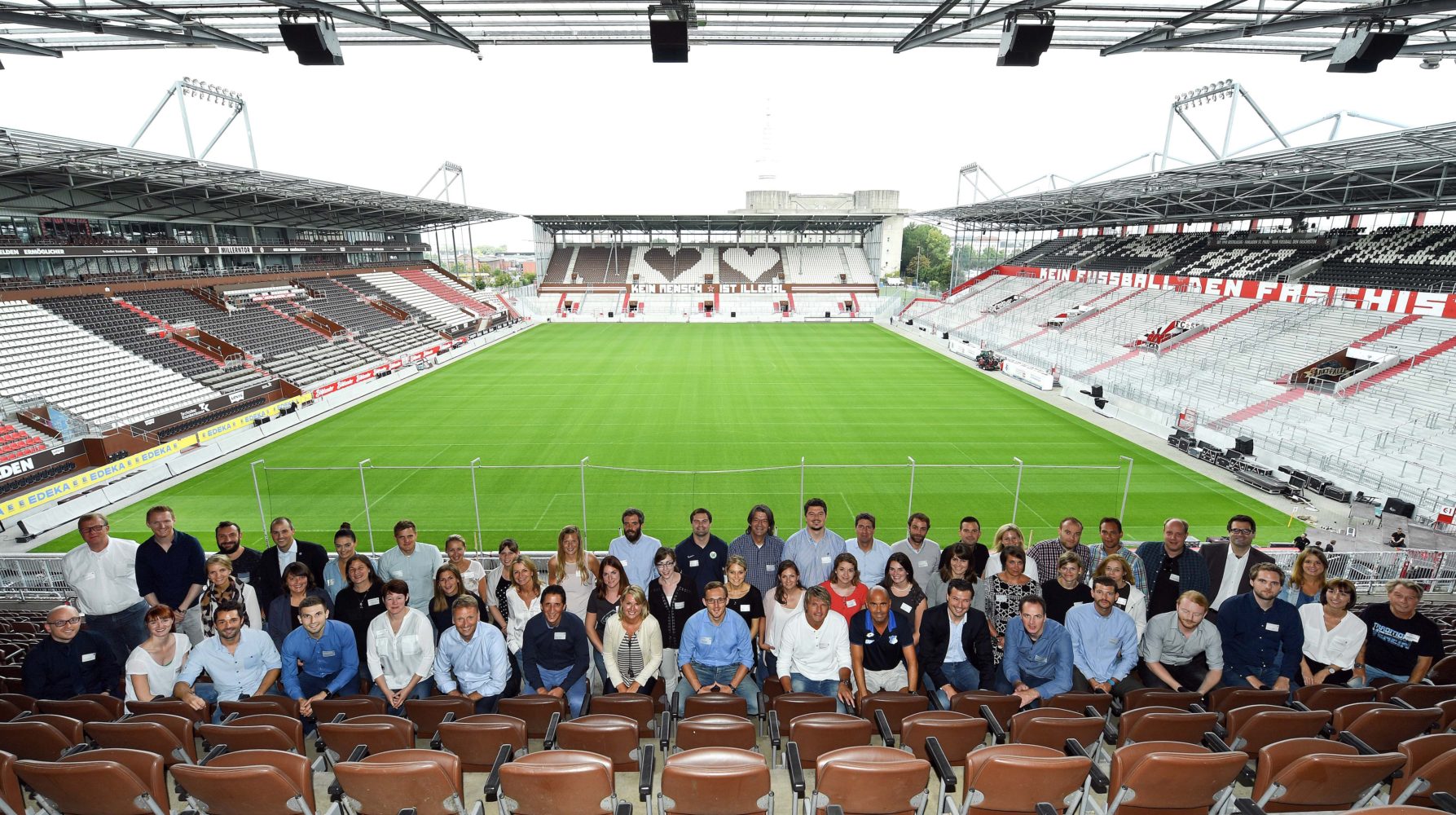 Eine große Gruppe von Menschen steht auf den leeren Sitzen eines Fußballstadions und blickt in die grüne Spielstätte. Im Hintergrund sind die leeren Tribünen und der Rasen zu sehen.