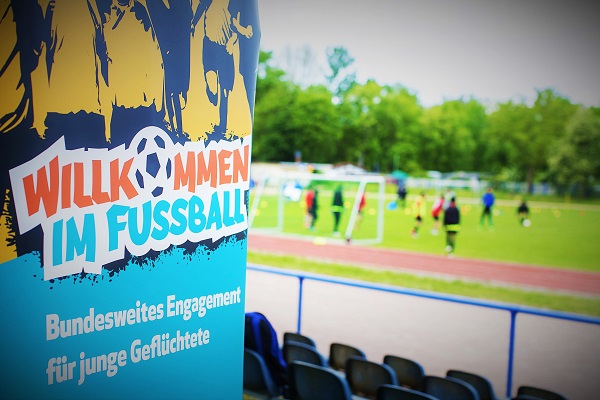 Im Vordergrund sieht man ein Plakat von "Willkommen im Fußball". Im Hintergrund sieht man viele Menschen bei einem Fußballspiel auf einem Fußballplatz.