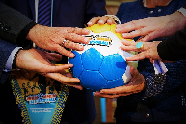 Eine Gruppe von Händen umfängt einen Fußball mit der Aufschrift "Willkommen im Fußball". Im Hintergrund ist ein Plakat mit demselben Aufdruck zu sehen. Das Bild symbolisiert die Unterstützung und das Engagement der DFL Stiftung für die Integration von Menschen durch Fußball.