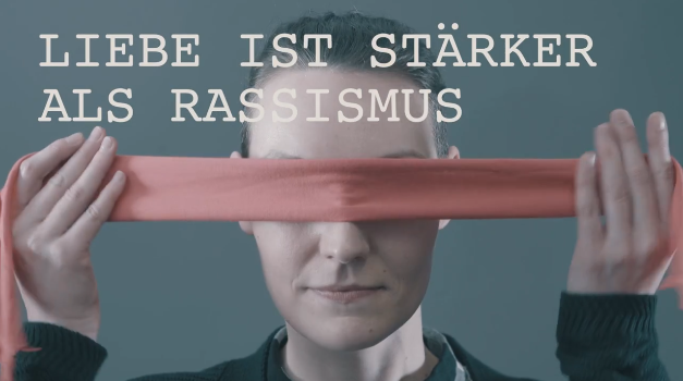 Ein Mann, vermutlich Jérôme Boateng, blickt ernst in die Kamera. Vor seinem Gesicht spannt sich ein rotes Band, das den Satz "LIEBE IST STÄRKER ALS RASSISMUS" überdeckt. Das Bild ist Teil einer Kampagne gegen Rassismus.