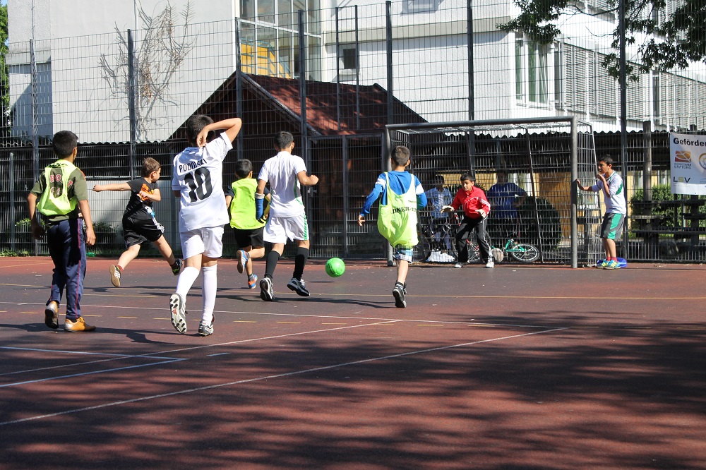Eine Gruppe von Jugendlichen spielt auf einem roten Bolzplatz Fußball. Ein Spieler in weißem Trikot mit der Nummer 10 schießt auf das Tor, während andere Spieler um ihn herum stehen und zum Angriff bereit sind. Im Hintergrund ist ein Junge im Rollstuhl zu sehen, der das Spiel verfolgt.