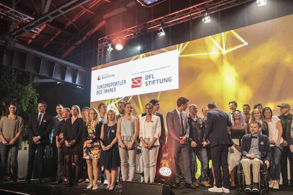 Eine Gruppe von jungen Athleten steht auf einer Bühne vor einem Banner mit der Aufschrift "Juniorsportler des Jahres" und dem Logo der DFL Stiftung. Einige der Athleten sitzen im Rollstuhl. Im Hintergrund sind Scheinwerfer und ein großes Banner zu sehen.