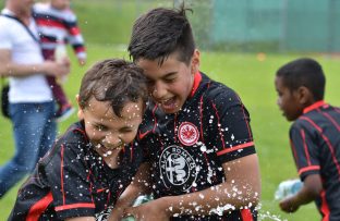 Zwei jubelnde Fußballspieler, beide tragen rote Trikots, werden mit Wasser übergossen. Im Hintergrund ist ein weiteres Kind zu sehen.