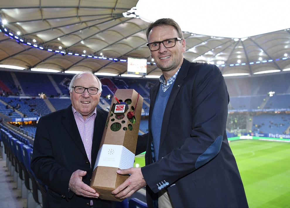 Zwei Männer überreichen sich eine geschenkt verpackte Spende im Fußballstadion. Links ein älterer Herr mit Brille und dunkelblauem Blazer, rechts ein Mann mit Hemd und dunkelblauem Blazer. Im Hintergrund sind die leeren Zuschauertribünen und der Rasen eines Fußballstadions zu sehen.