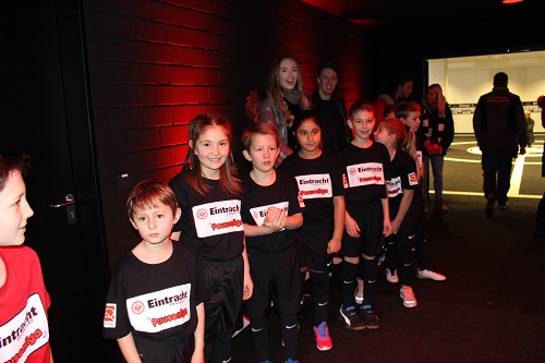 Eine Gruppe von Kindern in T-Shirts mit dem Aufdruck "Eintracht Frankfurt Pausenliga" steht in einem dunklen Gang und schaut in die Kamera. Einige Kinder halten kleine rote Bälle in den Händen. Im Hintergrund sind weitere Personen erkennbar.