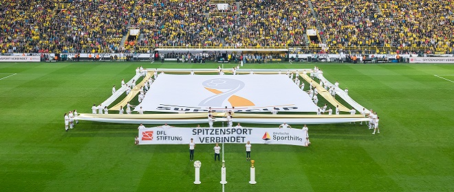 DFL Stiftung präsentiert eine große Bannerinstallation mit dem Schriftzug "SPITZENSPORT VERBINDET" auf dem Rasen des Signal Iduna Parks in Dortmund, umrahmt von Personen mit DFL-Logo und Sportgeräten, im Hintergrund ein voll besetztes Stadion.