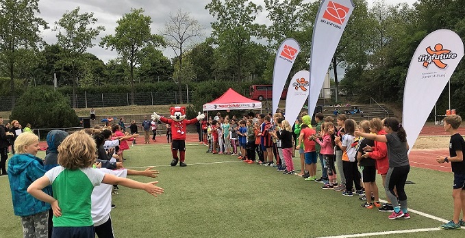 Eine Gruppe von Kindern und Jugendlichen nimmt an einer sportlichen Aktivität im Rahmen des "fit4future"-Programms der DFL Stiftung teil. Im Hintergrund sind Banner und ein rotes Zelt zu sehen.