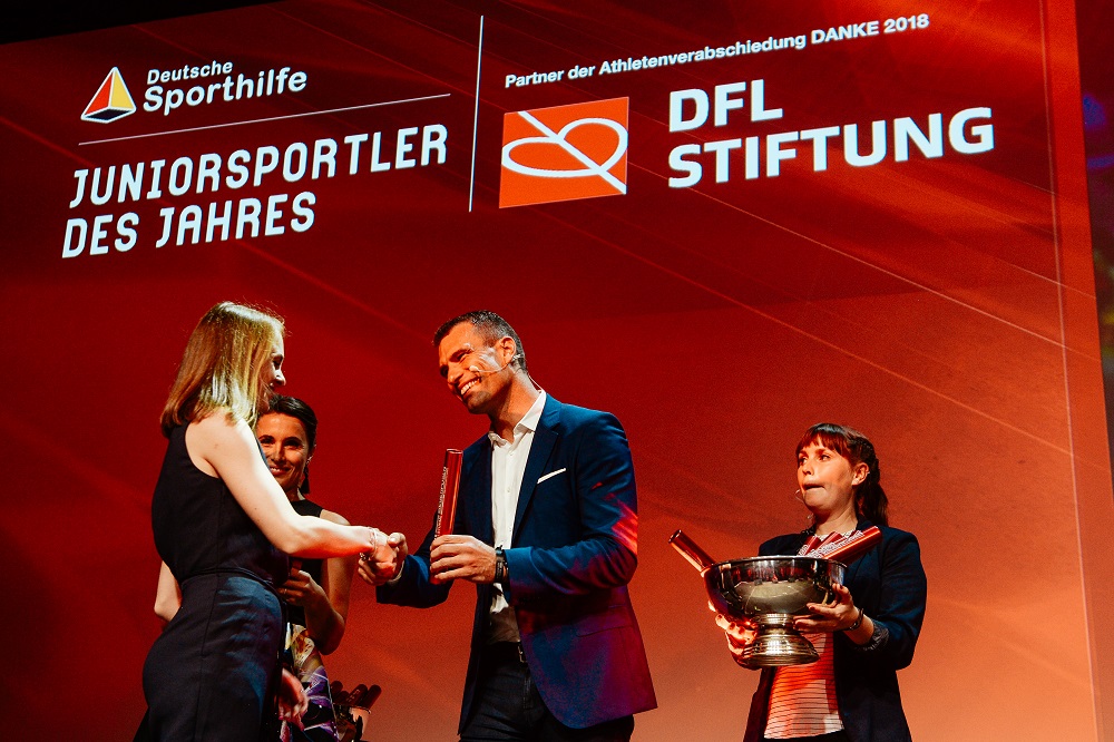 Lea-Jasmin Riecke, Weitspringerin, nimmt von einem Mann eine Auszeichnung als Juniorsportlerin des Jahres entgegen. Im Hintergrund eine weitere Frau mit einer Pokale. Auf dem roten Hintergrund des Bühnenbilds sind das Logo der Deutschen Sporthilfe und der DFL Stiftung zu sehen.