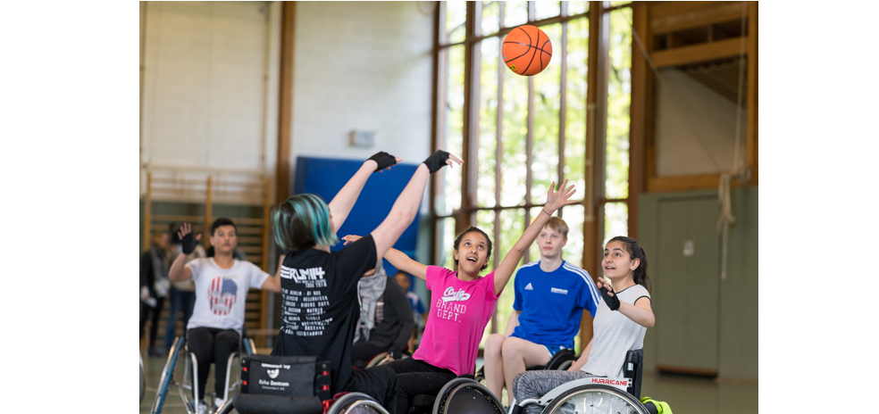 Rollstuhlbasketball: Eine Gruppe von Jugendlichen spielt in einer Sporthalle. Ein Mädchen in pinkem T-Shirt und blauem Helm springt in der Luft, um den Ball zu fangen. Im Hintergrund weitere Kinder im Rollstuhl.