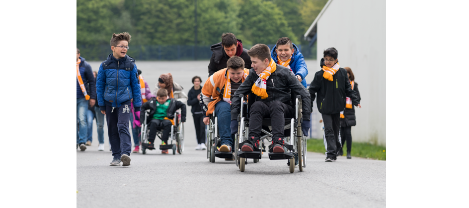 Eine Gruppe von Jugendlichen, einige in Rollstühlen, nimmt an einer Veranstaltung im Freien teil. Sie tragen orangefarbene Schals und bewegen sich auf einem Weg entlang. Im Hintergrund sind weitere Personen und grüne Bäume zu sehen.
