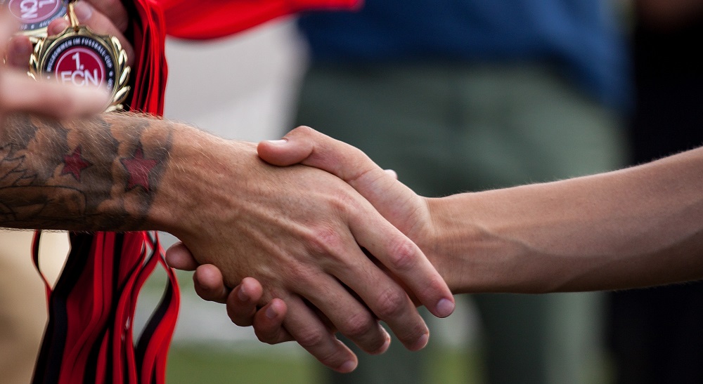 Nahaufnahme von zwei Händen, die sich den Handschlag geben. Im Hintergrund ist ein rotes Trikot mit dem Logo des 1. FC Nürnberg zu sehen und eine grüne Rasenfläche.