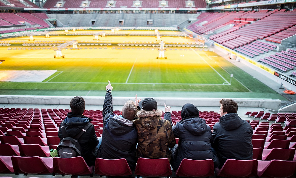Eine Gruppe Jugendlicher sitzt auf roten Stadiontribünen und blickt auf das grüne Fußballfeld.