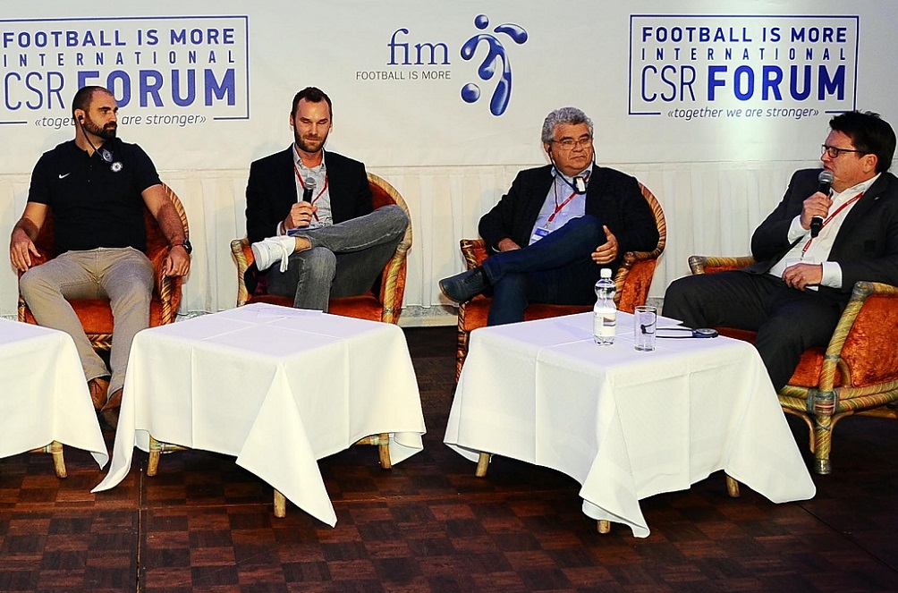 Vier Männer sitzen an einem Tisch auf einem Podium und nehmen an einer Diskussion teil. Im Hintergrund ist ein Banner mit der Aufschrift "FOOTBALL IS MORE INTERNATIONAL CSR FORUM" zu sehen.