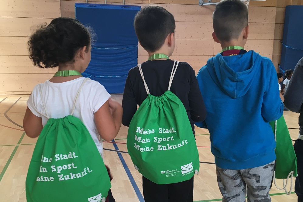 Three children (one girl and two boys) are standing in a gym with their backs to the camera. Two of the children are carrying gym bags with the inscription "My town. My sport. My future.".