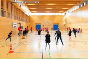 Eine Gruppe von Kindern spielt in einer großen Sporthalle. Im Hintergrund ist eine Wand mit einem Basketballkorb und ein Banner mit dem Logo von Eintracht Frankfurt zu sehen. Einige Kinder werfen und fangen Bälle, andere stehen und beobachten das Spiel. Die Sporthalle ist hell und gut beleuchtet.