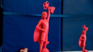 Eine Großaufnahme des Siegerpokals des Programms step kickt!. Der Pokal ist rot und ist eine Figur, die ein Superhelden-Cape umhat und einen kleinen Pokal in die Höhe streckt.