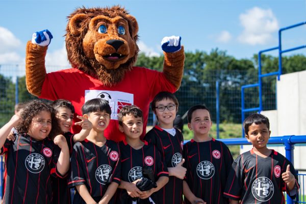 Sieben Jungs in schwarzen Trikots von Eintracht Frankfurt stehen vor Litti, dem Maskottchen von "Fußball trifft Kultur". Litti ist ein Löwe mit rotem T-Shirt.