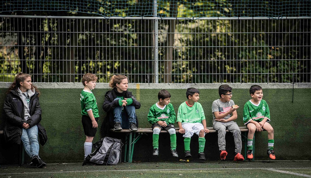 Junge Fußballspielerinnen und Fußballspieler des Bremer „Willkommen im Fußball“-Bündnisses sitzen auf den Bänken neben dem Fußballfeld und schauen das Spiel zu.