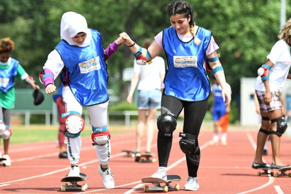 Zwei junge Frauen mit Migrationshintergrund fahren Skateboard. Dabei halten sie sich an jeweils einer Hand fest.