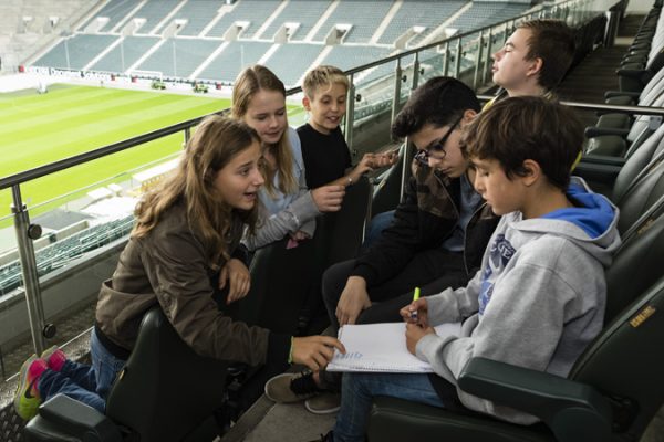 Eine Gruppe Kinder und Jugendlicher Teilnehmender des Programms Lernort Stadion sitzt auf den leeren Tribünen eines Fußballstadions. Ein Mädchen zeigt einem jungen etwas auf einem Blatt Papier, dass dieser in der Hand hält.