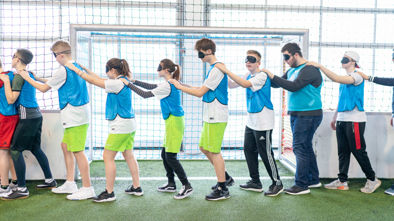 Young participants in the Lernort Stadion program stand in a row behind each other and hold the person in front of them by the shoulders with their hands. They wear eye covers to experience how visually impaired people feel.