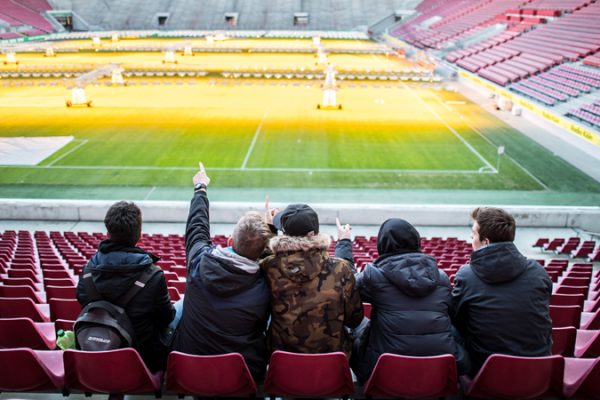 A small group of students sits together in an empty soccer stadium and looks out onto the field from the stands. The group is taking part in the DFL Foundation's "Lernort Stadion" program.