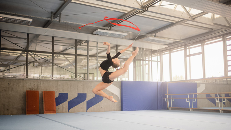 Rhythmic gymnast Noemi Peschel demonstrates a jump in a gymnasium. She holds a red swing band in her hand.