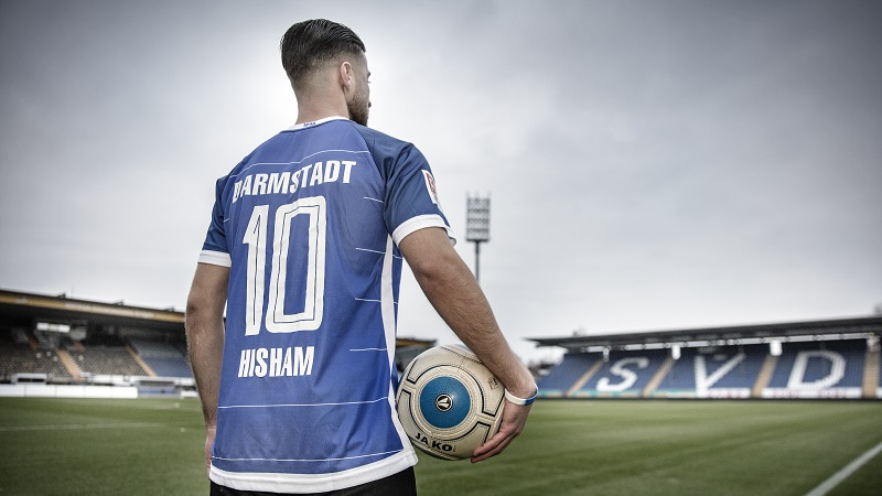 Ein junger Mann steht mit einem Ball unter dem Arm im Stadion des SV Darmstadt 98. Er trägt ein blaues Trikot von Darmstadt 98 mit der Rückennummer 10.