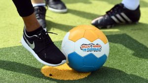 Ein Fußball mit der Aufschrift "Willkommen im Fußball" steht auf einem grünen Fußballrasen. Im Hintergrund sind die Füße von Kindern oder Jugendlichen zu sehen.