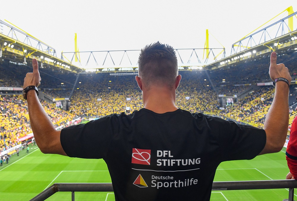 Ein Mann im schwarzen T-Shirt mit Logos von der DFL Stiftung und Deutschen Sporthilfeguckt auf das Stadion und zeigt beide Daumen nach oben