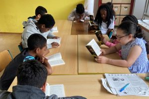 Konzentrierte Kinder sitzen an Tischen und lesen oder arbeiten mit Büchern und Arbeitsblättern. Im Hintergrund stehen Regale mit weiteren Materialien.