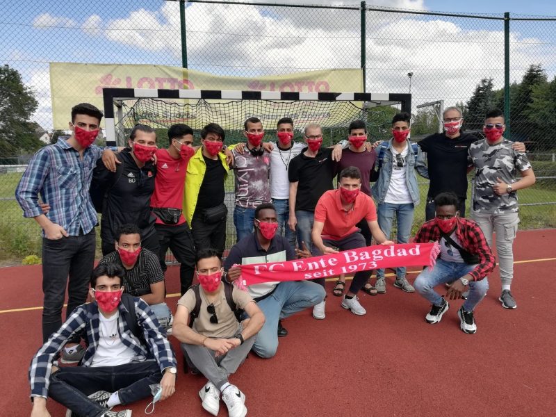 Junge Geflüchtete, des FC Ente stehen mit Masken vor dem Fußballtor und zeigen einen roten Schal mit dem Namen des Clubs