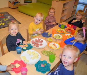 Mehrere Kleinkinder sitzen an einem mit bunten Tellern und verschiedenen gesunden Lebensmitteln gedeckten Tisch und lachen in die Kamera.