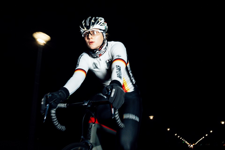 Lea Sophie Friedrich fährt im Dunkeln mit ihrem Rennrad. Sie trägt dabei einen weißen Helm und ein Trikot der deutschen Rad-Nationalmannschaft. Auf dem weißen Shirt befinden sich der Bundesadler im Brustbereich sowie Streifen in den Farben der Deutschland-Flagge auf Höhe der Oberarme und auf der Vorderseite des Trikots.