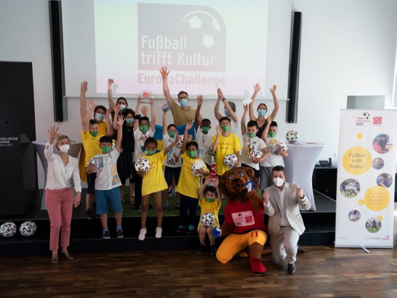 Schüler der Karmeliterschule winken mit den Jurymitgliedern und der Maskotte des Programmes ,,Fußball trifft Kultur"