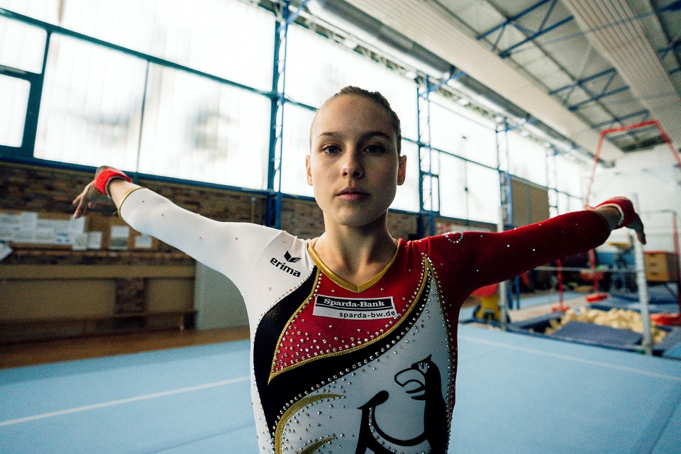 Sarah Voss steht in einer Turnhalle und trägt dabei einen Turnanzug, auf dem auf der Vorderseite der Bundesadler großflächig abgebildet ist. Voss schaut in die Kamera und streckt ihre beiden Arme jeweils zur Seite weg.