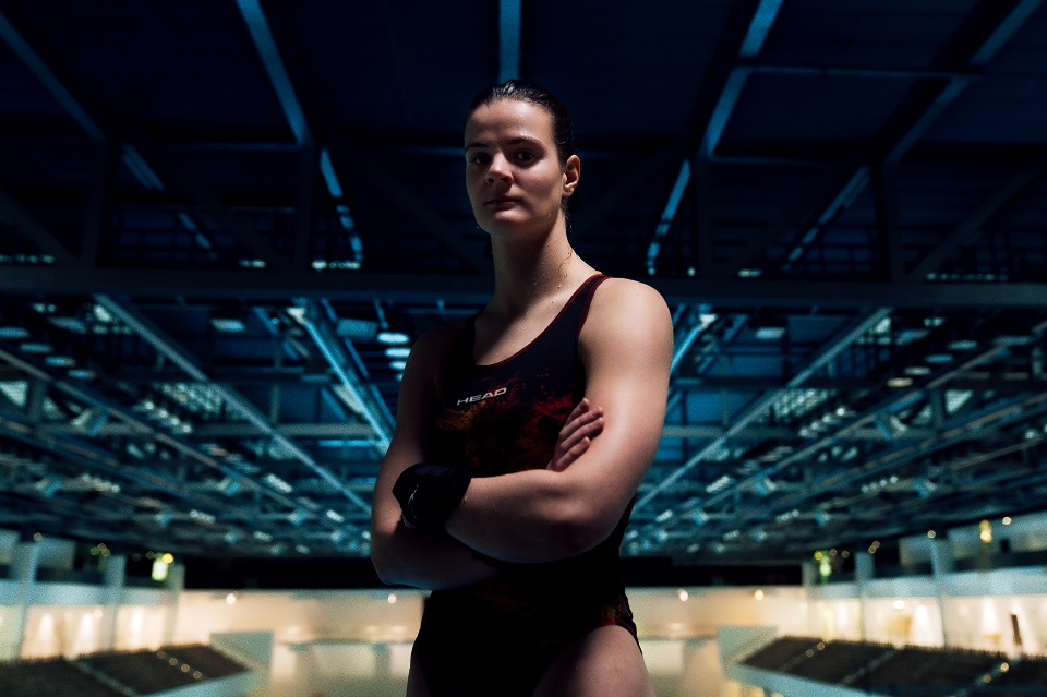 Elena Wassen steht in einem Badeanzug bekleidet mit verschränkten Armen auf einer Sprungplattform in einer Schwimmhalle. Wassen steht mit dem Rücken zum Wasser und schaut dabei in die Kamera.