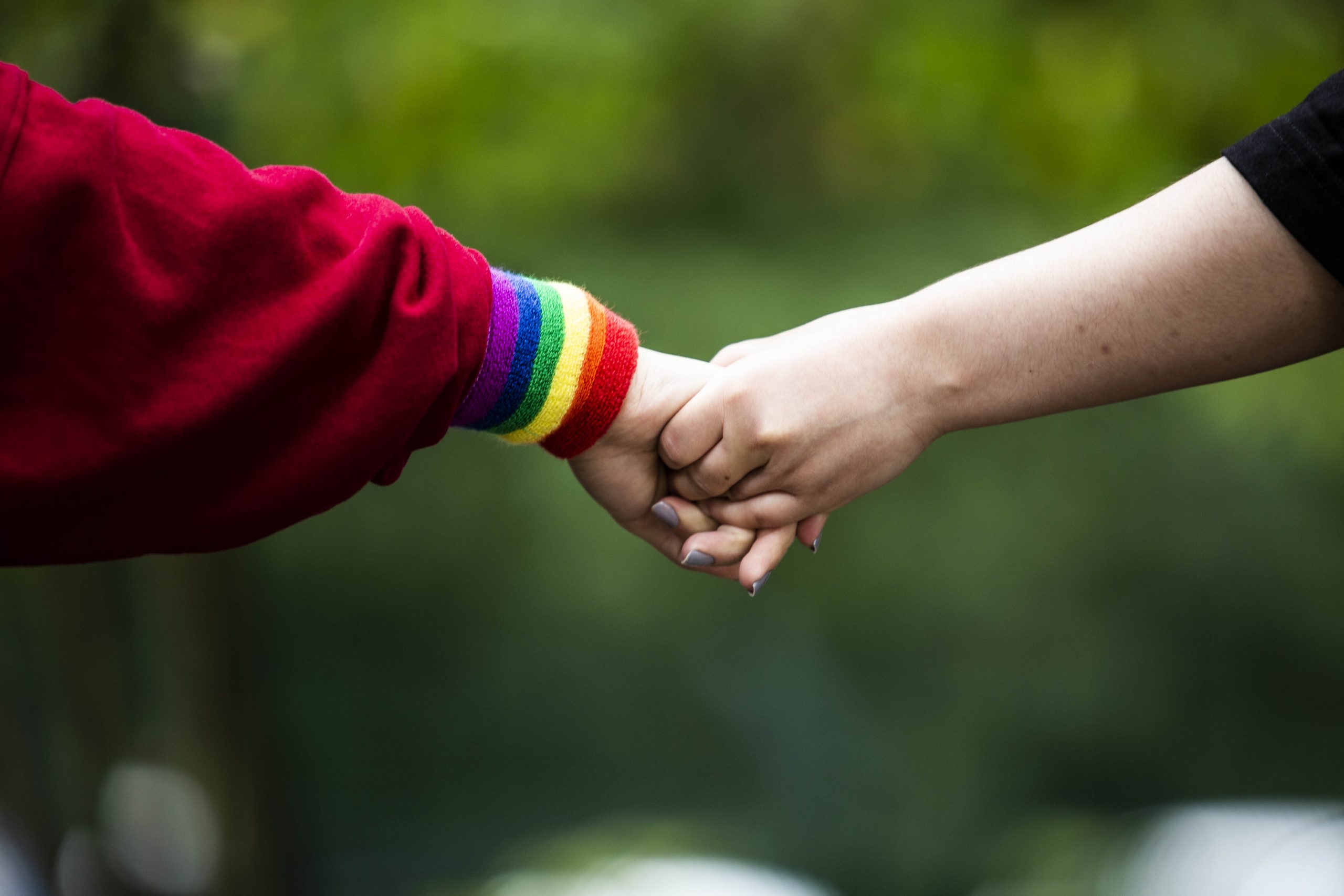 Zu sehen sind zwei sich haltende Hände. An einem Handgelenk ist ein Schweißband in Regenbogenfarben zu sehen.