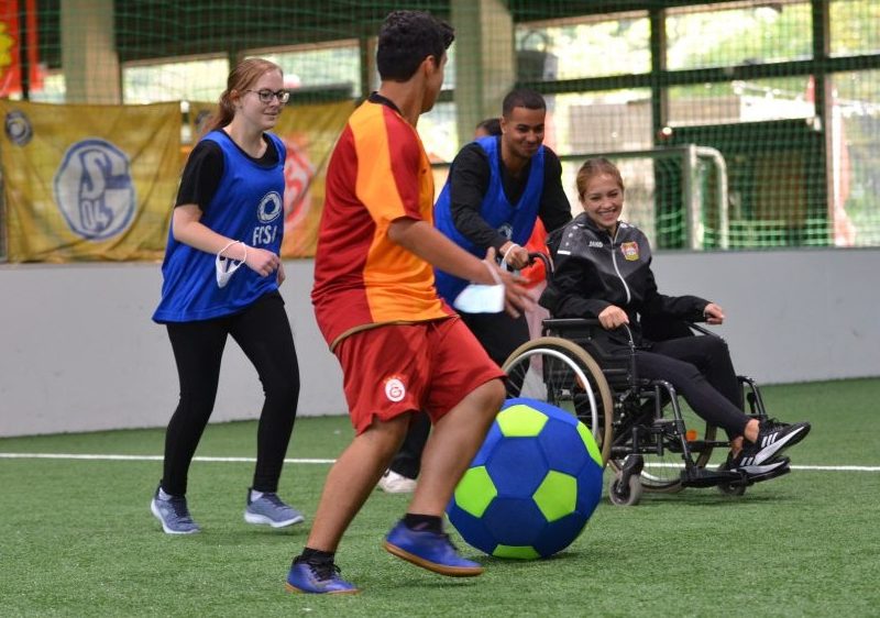 Ein Junge schiebt das Mädchen im Rollstuhl und beide spielen Fußball mit zwei weiteren Teilnehmenden der Coach-Ausbildung in einer Fußballhalle.