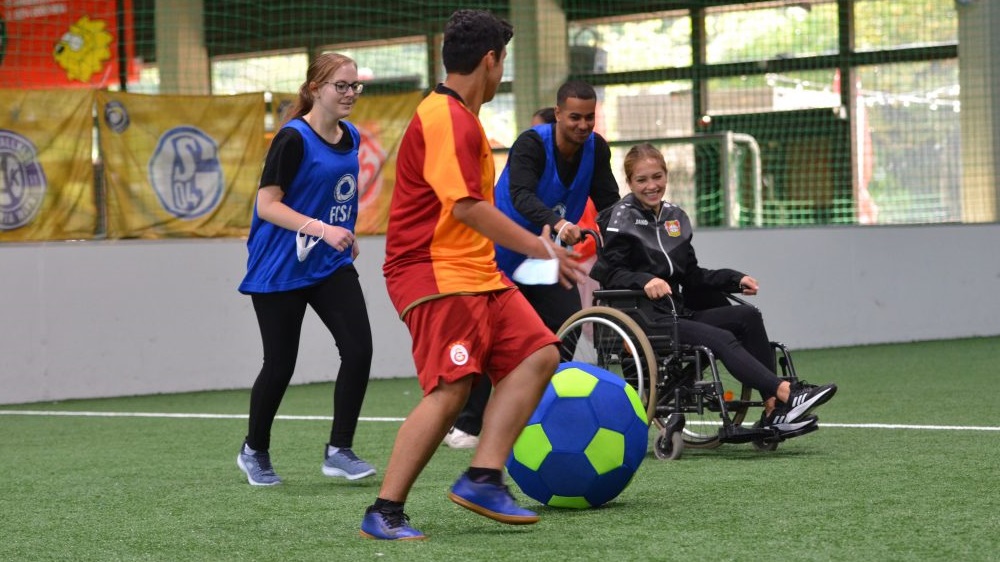 Ein Junge schiebt das Mädchen im Rollstuhl und beide spielen Fußball mit zwei weiteren Teilnehmenden der Coach-Ausbildung in einer Fußballhalle.