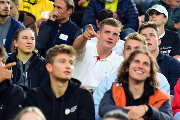 Young people are sitting in the stands at the soccer stadium. One of them points his finger at something.