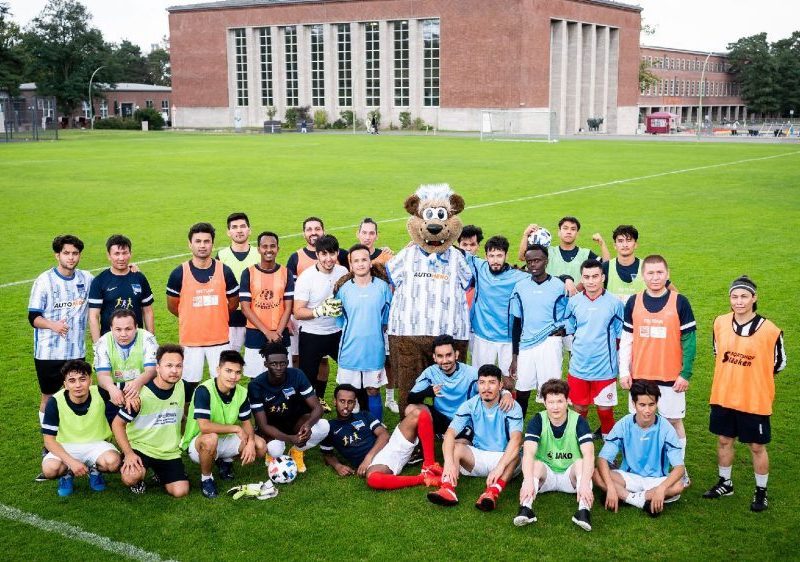 Die Teilnehmende des Abschlussturniers stehen mit der Maskotte auf dem Fußballfeld.