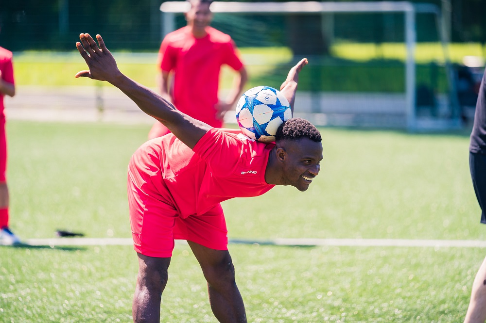 Ein Fußballspieler im roten Trikot beugt sich nach vorne und balanciert auf dem oberen Rücken einen Fußball.