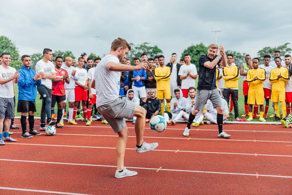 Zwei Männer machen auf einer Tartanbahn Fußballtricks. Um sie herum stehen Teilnehmende von „Willkommen im Fußball"