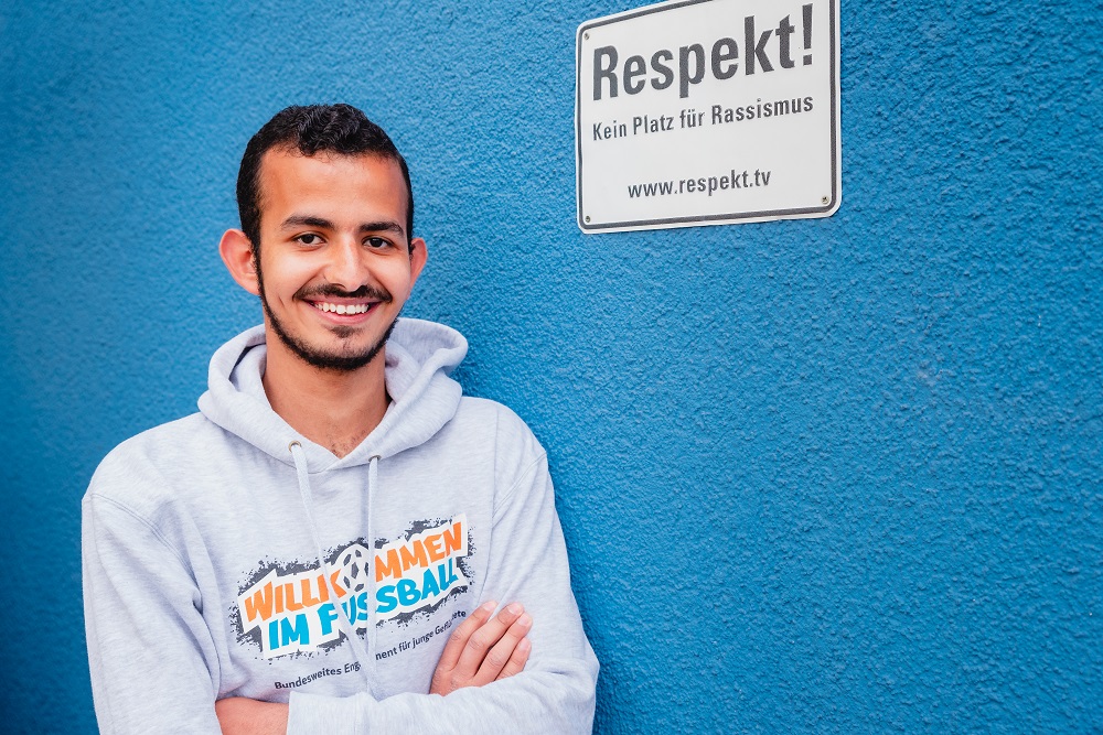 Saleh steht mit den Armen vor der Brust verschränkt vor einer blauen Wand. An der Wand hängt ein Schild, auf dem "Respekt" steht.