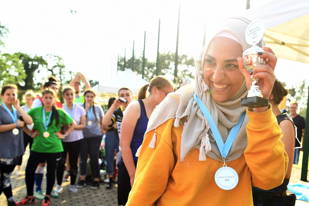 Im Vordergrund ist Marah zu sehen. Sie trägt einen orangefarbenen Pulli und hat eine Medaille um den Hals. Im Hintergrund sind weitere Menschen.