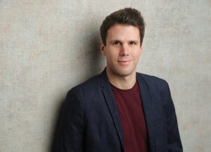 Moritz Glenk, Referent der DFL Stiftung, im Porträt. Er trägt ein rotes Shirt und eine dunkle Jacke und lächelt in die Kamera.